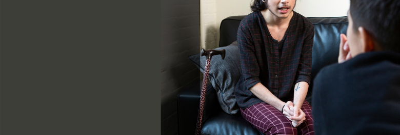 A genderqueer person sits on a leather couch with their hands clasped. Their cane rests against the couch beside them. Their face is cropped out just above their nose. They are speaking with someone in the foreground facing away from the viewer and leaning forward to listen.