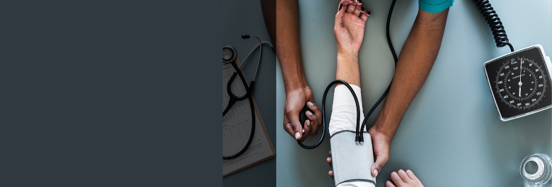 From overhead a Black health provider takes the blood pressure of another tanned skinned individual. A clipboard and stethoscope is also pictured to portray a health setting.